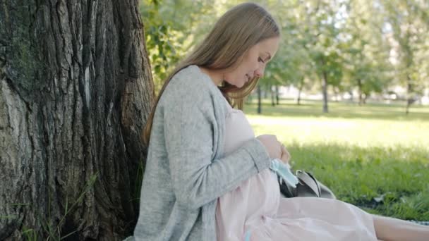 Söt gravid kvinna som har kul med baby skor röra magen ler sitter på gräsmattan i parken — Stockvideo