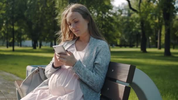 Atraente jovem senhora futura mãe tocando tela de telefone inteligente e sorrindo relaxante no banco no parque da cidade — Vídeo de Stock