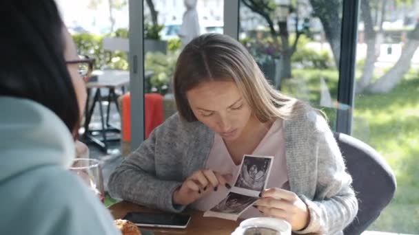 Mulher grávida alegre compartilhando imagem de ultrassonografia do bebê com amigos sentados à mesa no café — Vídeo de Stock