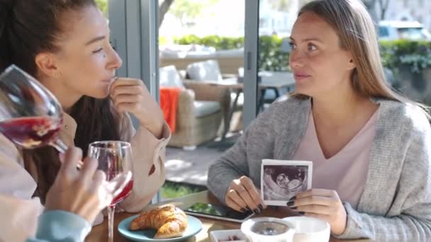 Esperando mãe mostrando imagem de ultra-som para amigos do sexo feminino desfrutando de dia de verão no café — Vídeo de Stock