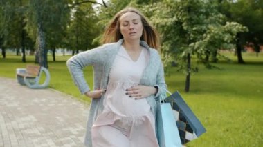Parkta elinde poşetlerle dolaşıp sırt ağrısı çeken hamile bir kadın.