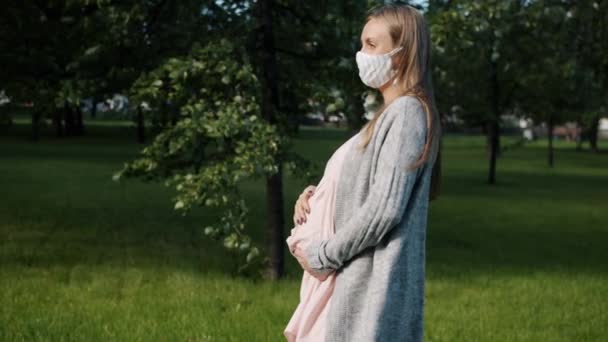 Joven embarazada parada al aire libre en el parque de la ciudad usando mascarilla médica durante la pandemia — Vídeos de Stock