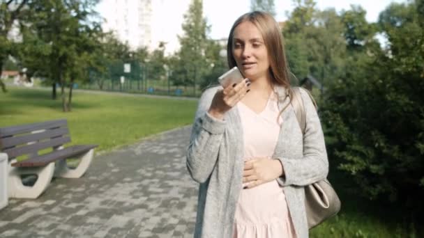 Chica embarazada alegre caminando en el parque de la ciudad enviando y escuchando mensajes de voz en el teléfono inteligente — Vídeo de stock