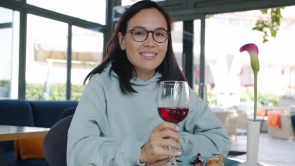 Portret van een mooie jonge brunette aan tafel in café met een glas wijn en een glimlachend dessert — Stockvideo