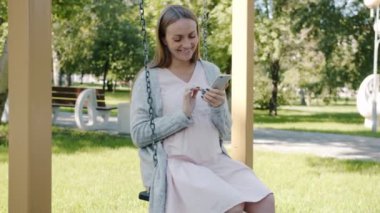 Şehir parkında salıncakta sallanan ve akıllı telefon tebessümü kullanan mutlu hamile kadın.