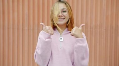 Neşeli sarışın kız iki eliyle baş parmak işareti yapıyor ve açık havada gülümsüyor.