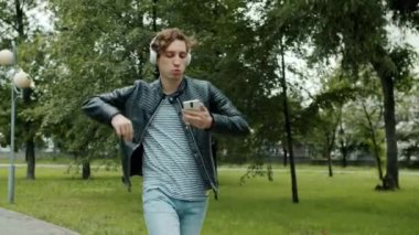 Şehir parkında tek başına dans eden, kulaklık takan ve akıllı telefon kullanan neşeli bir adam.