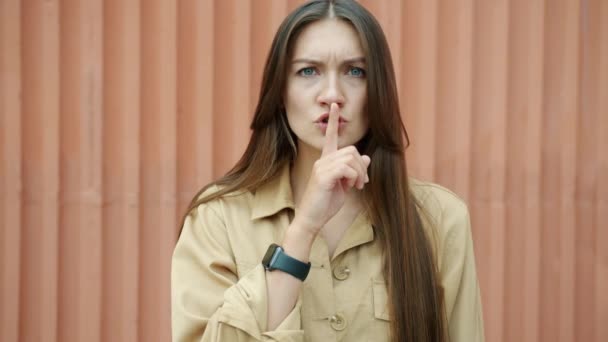 Slow motion of serious young woman touching lips with finger asking for silence looking at camera standing outdoors — Stock Video