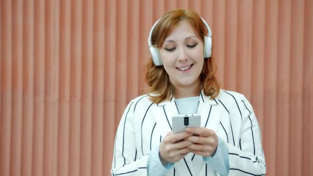 Slow motion portret van schattige roodharige vrouw met een koptelefoon met behulp van een smartphone en dansen buiten — Stockvideo