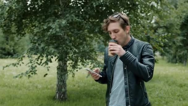 Cámara lenta de chico atractivo usando teléfono inteligente y beber para ir a caminar café al aire libre en el parque de la ciudad — Vídeo de stock