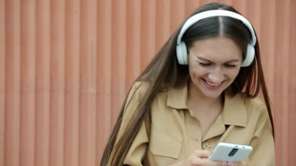 Akıllı telefon kullanan, kulaklıkla müzik dinleyen ve dışarıda dans eden neşeli genç bir kadın. — Stok video
