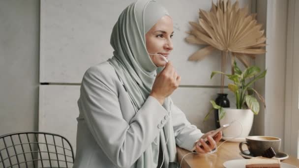 Chica hijabi feliz hablando en el teléfono móvil con auriculares celebración de teléfono inteligente en la cafetería moderna — Vídeos de Stock