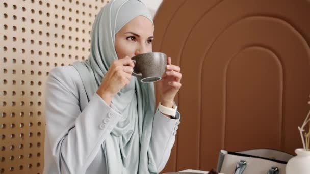 Retrato de hijabi joven pensativo disfrutando de una taza de café y sonriendo en el interior de la cafetería — Vídeos de Stock