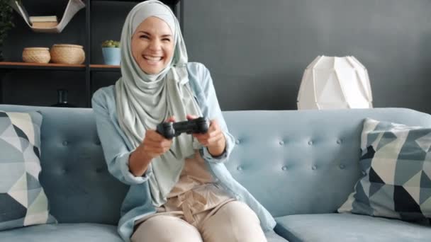 Ritratto di allegra giovane muslimah che gioca al videogioco divertendosi e ridendo in casa — Video Stock