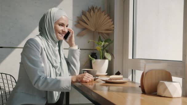 Mujer árabe alegre vistiendo hijab hablando por celular y sonriendo disfrutando del día en la cafetería — Vídeo de stock