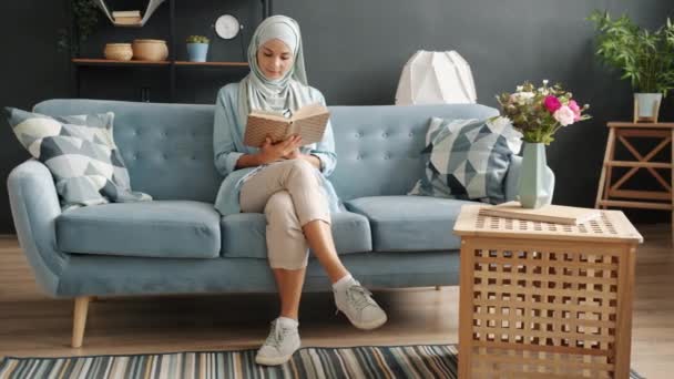Vrolijk meisje dragen hijab lezen boek en glimlachen genieten van vrije tijd in appartement — Stockvideo