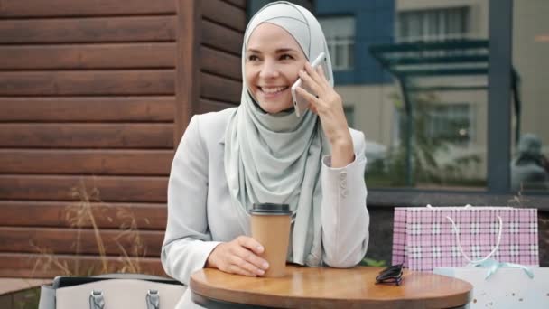 Mujer musulmana despreocupada vistiendo el tradicional hijab charlando en el teléfono móvil sonriendo en la cafetería al aire libre — Vídeo de stock