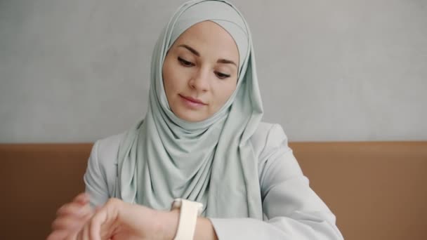 Vrolijke moderne muslimah kijken naar slimme horloge scherm en glimlachen binnen in cafe — Stockvideo