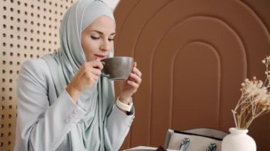 Kafede kahve içen, sonra akıllı saati kontrol eden ve kafasını sallayan ciddi bir muslimah portresi.