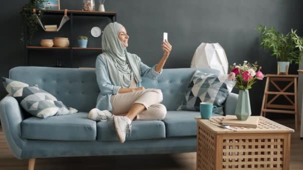 Gülümseyen genç Müslüman kadın güzel bir dairedeki akıllı telefon ekranına bakarak online video görüşmesi yapıyor. — Stok video