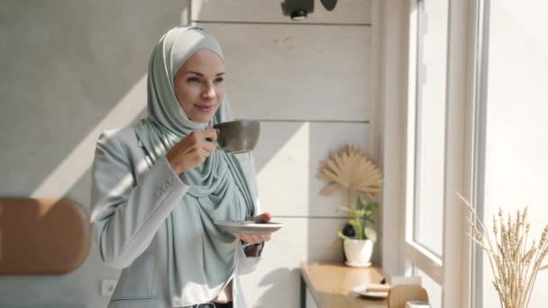 Lambat gerak bahagia ras wanita mengenakan hijab minum kopi dan melihat kota melalui jendela di kafe — Stok Video