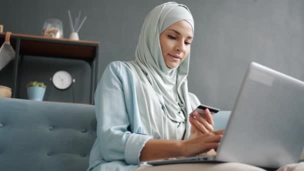 Fröhliches muslimisches Mädchen im Hijab shoppt online mit Bankkarte und Laptop von zu Hause aus — Stockvideo