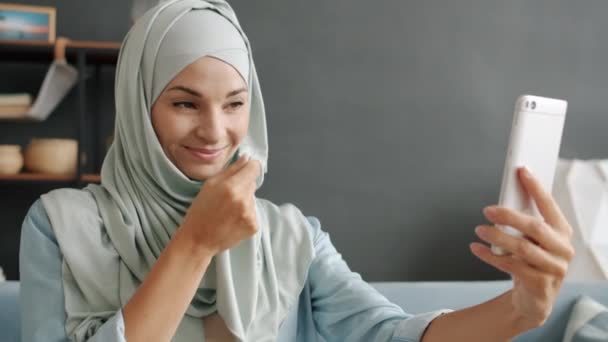 Медленный портрет веселой мусульманки, позирующей перед камерой смартфона, делающей селфи в помещении — стоковое видео