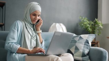 Neşeli iş kadını cep telefonuyla sohbet ediyor ve dairede dizüstü bilgisayarla çalışıyor.