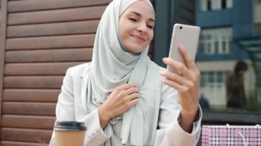 Neşeli muslimah dışarıdaki kafede akıllı telefon kamerasıyla selfie çekiyor.