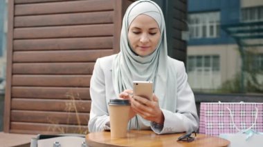 Tesettürlü, akıllı telefon kullanan ve açık kafede kahve içen kaygısız bir kızın portresi.