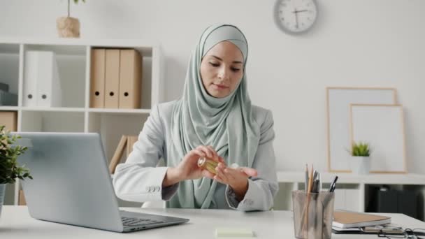 Portret van een moslim vrouw die naar kantoor komt, handen reinigt met gel en dan aan het bureau werkt. — Stockvideo