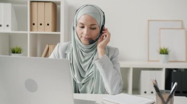 Türbanlı modern muslimah çağrı merkezinde çalışan bir müşteriyle mikrofonlu kulaklıklar kullanarak konuşuyor.