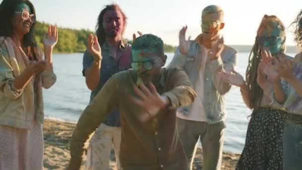 Fröhlicher Mann aus dem Nahen Osten, der mit Gullyfarbe bedeckt ist und mit Freunden draußen auf einer Strandparty tanzt — Stockvideo
