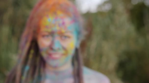 Zoom-in portrait of beautiful girl with dreadlocks and colorful face smiling standing outdoors at Holi party — Stock Video