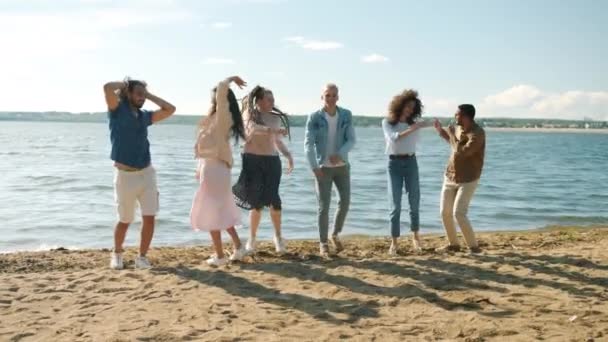 Retrato en cámara lenta de alegres millennials bailando en la playa y luego saltando levantando los brazos riendo — Vídeos de Stock