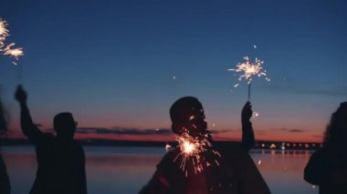 Gece kumsalında dans eden insan siluetinin yavaş çekimleri Bengal ışıkları sallayarak etkinliğin tadını çıkarıyor.