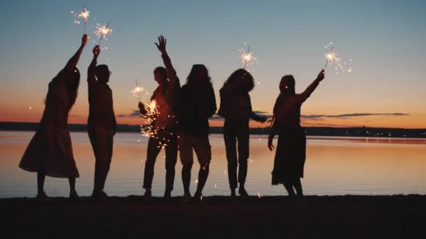 Lento movimiento de siluetas masculinas y femeninas bailando en la playa sosteniendo luces de bengala por la noche — Vídeos de Stock