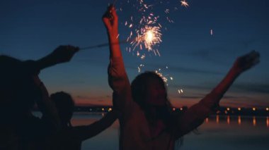 Havai fişekli mutlu öğrenciler karanlık sahilde dans ediyor. Gece partisinin tadını çıkarıyorlar.