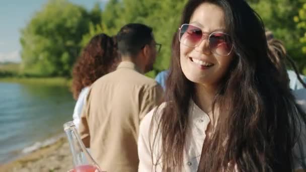 Retrato de menina asiática feliz vestindo óculos de sol dançando na praia segurando garrafa com bebida — Vídeo de Stock