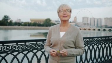 Nehir kenarında mutlu bir gülümsemeyle yürüyen olgun bir kadının yavaş çekim portresi.