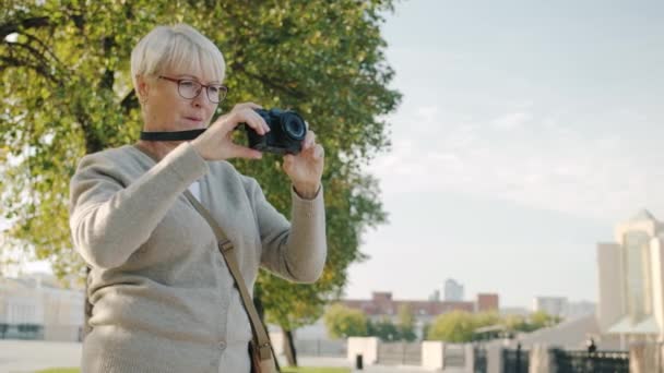 Gammal kvinnlig fotograf tar bilder med modern kamera i vacker sommarstad — Stockvideo
