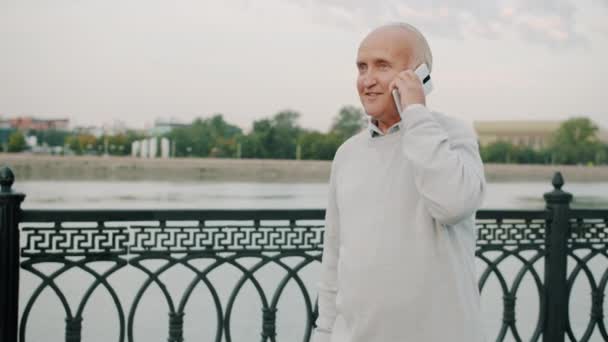 Vidám idős férfi beszél mobilon séta a folyóparton élvezi a beszélgetést — Stock videók