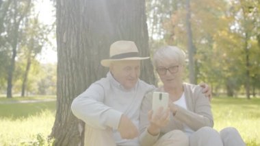 Neşeli yaşlı çift akıllı telefon ekranına bakıyor güneşli bir günde parkta eğleniyorlar.