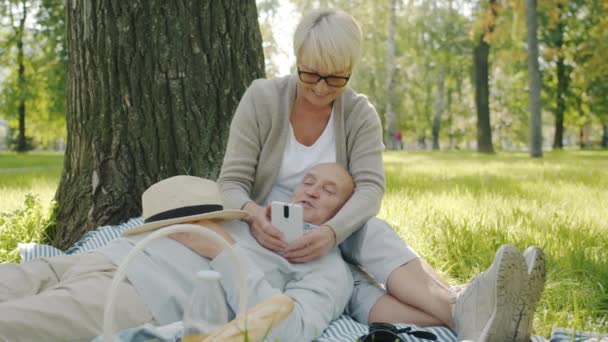 Старые муж и жена с помощью смартфона болтая расслабляясь на траве в парке — стоковое видео