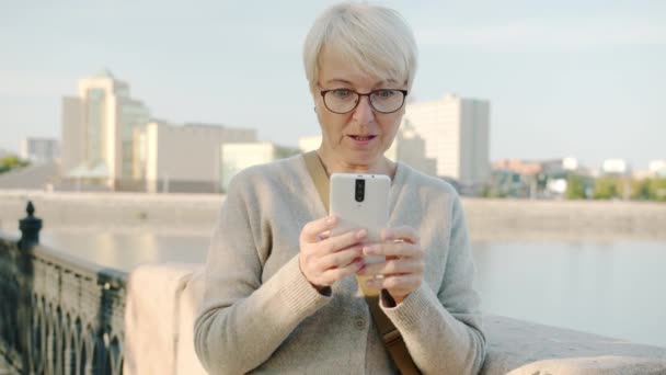 Lambat gerak wanita muda SMS dengan smartphone tersenyum menikmati kabar baik berdiri di tanggul sungai — Stok Video