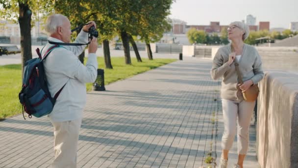 Neşeli olgun kadın fotoğraf makinesi için poz verirken kocası arka planda modern şehirle fotoğraf çekiyor. — Stok video