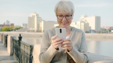 Şehirde sosyal medyadan keyif alan akıllı telefon kullanan mutlu yaşlı kadın portresi