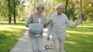 Mutlu son sınıf çifti yaz günü pikniğe hazır yiyecek ve battaniyeyle parkta yürüyor.