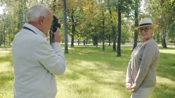 Slow motion av senior dam poserar för kameran medan make tar bilder med modern enhet i parken — Stockvideo