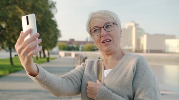 Senhora sênior bonita levando selfie com câmera de telefone inteligente posando no dique do rio no dia de verão — Vídeo de Stock